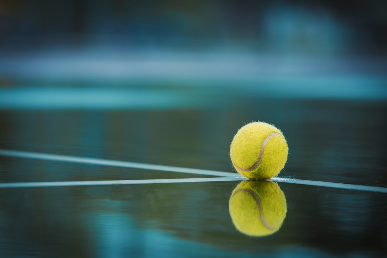 services-04 A tennis ball sits on a wet court with its reflection visible, capturing a serene sports moment.